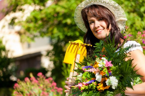 A woman with dark brown, wavy hair wearing a straw hat and a white top is standing in a well-maintained front garden. She is holding a bright yellow garden rake in one hand and a large, colorful bouquet of freshly cut flowers, including pink, white, orange, and purple blooms, along with lush green foliage in the other. Behind her, the garden features a neatly trimmed hedge lining a paved pathway, a lush green lawn with dense, healthy grass, and various trees and shrubs creating a vibrant, natural backdrop. The scene is bathed in natural daylight, indicating clear weather, and the garden is part of a residential property in Hammersmith, with soft-focus residential buildings visible in the background. The setting exemplifies outdoor garden maintenance and floral arrangement activities typical of professional gardening services provided by Hedge Trimming Hammersmith, highlighting a tidy and picturesque outdoor space. The overall composition reflects a well-kept, colorful garden environment suitable for landscaping and outdoor care.