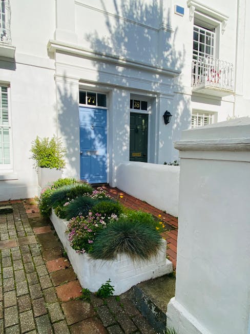 A small front garden area in front of a white residential building features a narrow brick pathway leading to a blue front door with the number 78 displayed above it. The garden bed is elevated with a